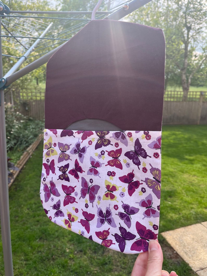 Pode incluir: Uma bolsa de tecido roxo com um forro de tecido branco com estampa de borboleta. A bolsa est&aacute; pendurada em um varal.