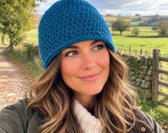 May include: A deep teal blue beanie with a textured, crocheted stitch pattern. The hat features a rounded crown and a simple, clean edge that fits snugly around the head, showcasing a consistent, dense yarn construction throughout the entire piece.