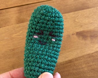 May include: Handmade green crochet pickle with embroidered facial features. The pickle has a smiling face with black eyes and pink cheeks. The crochet work is dense and textured, giving the pickle a three-dimensional appearance.