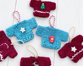 May include: A collection of six miniature knitted jumper ornaments in burgundy and teal. Each jumper features a hanging loop and a decorative wooden embellishment, including a green Christmas tree, a red mitten, and a grey reindeer.