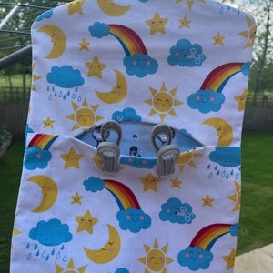 Puede incluir: Un organizador de colgar de tela blanca con un patr&oacute;n de arco&iacute;ris, nubes, estrellas, lunas y soles. El organizador tiene dos bolsillos y est&aacute; dise&ntilde;ado para colgar en una percha.