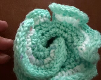 May include: A round, crocheted item featuring a spiral design in shades of mint green and white. The yarn texture is visible throughout the piece, which is constructed with a series of layered, ruffled loops that create a dense, circular form.