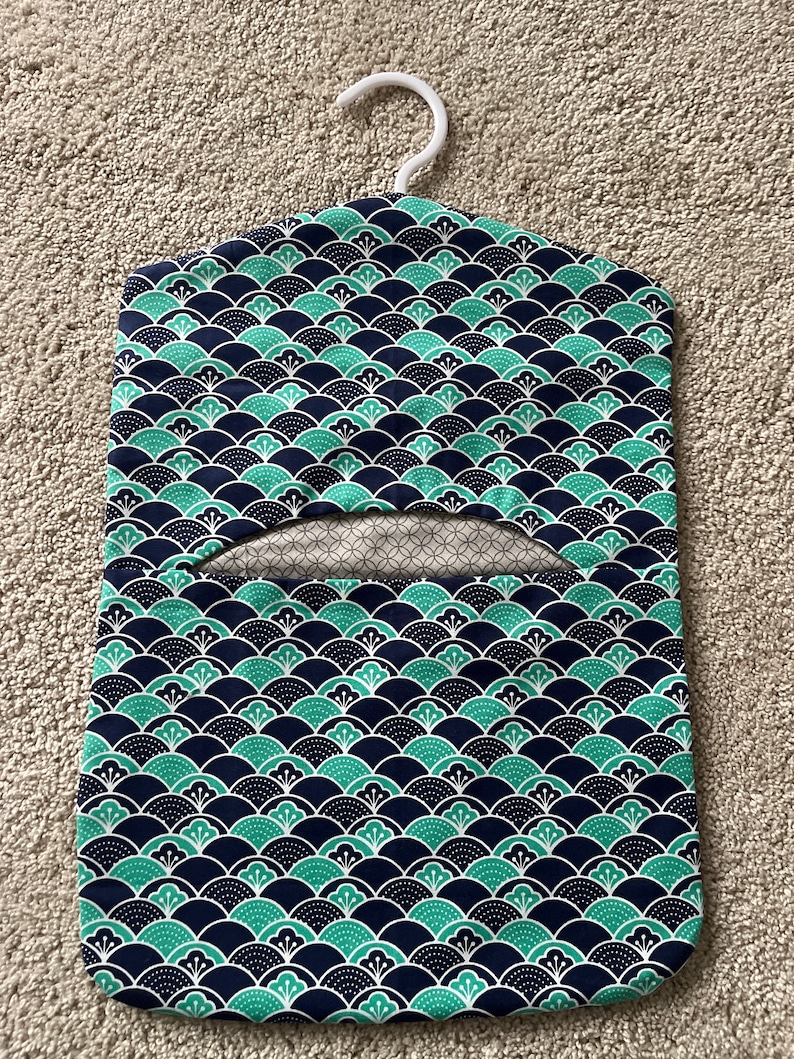 May include: A blue and green fabric garment bag with a white hanger. The bag has a repeating pattern of green and white stylised flowers on a dark blue background.