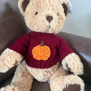 Pode incluir: Um urso de peluche com pelo castanho claro, interior das orelhas castanho escuro e almofadas nas patas castanho escuro. O urso veste uma camisola de malha bordeaux com uma aplicação de abóbora laranja texturizada no peito. Etiqueta branca.