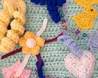 May include: A light mint green crochet sensory mat featuring various attached elements including a yellow ruffled circle, a blue spiral, a yellow bunny, a pink star with a wooden button, a white flower, a white heart, and colourful beads.