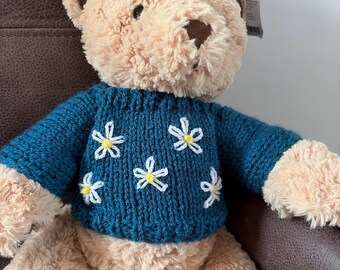 May include: A plush teddy bear with light tan fur, dark brown ears, and a dark brown nose. The bear is wearing a dark teal knitted jumper featuring a pattern of embroidered white flowers with yellow centres. A small white product tag is attached.