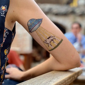 May include: A temporary tattoo of a cow being abducted by a UFO. The UFO is blue and white with a yellow beam of light shining down on the cow.