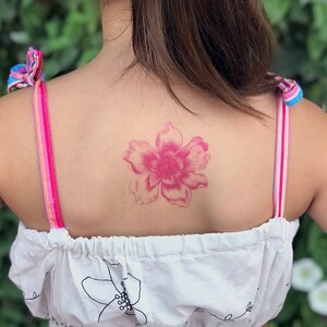 Puede incluir: Un tatuaje de una flor rosa en la espalda de una persona. La flor tiene cinco p&eacute;talos y est&aacute; en un estilo acuarela.