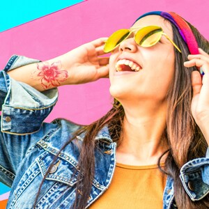 Puede incluir: Una mujer con el pelo largo y casta&ntilde;o lleva una chaqueta vaquera, una camisa amarilla y una diadema colorida. Lleva gafas de sol amarillas y sonr&iacute;e. Se puede ver un tatuaje de una flor rosa en su mu&ntilde;eca.