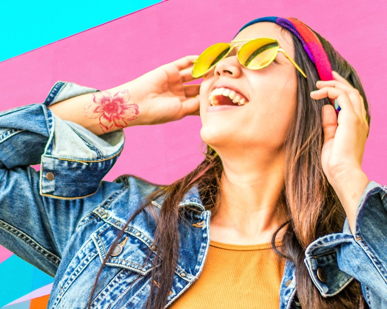 Puede incluir: Una mujer con el pelo largo y casta&ntilde;o lleva una chaqueta vaquera, una camisa amarilla y una diadema colorida. Lleva gafas de sol amarillas y sonr&iacute;e. Se puede ver un tatuaje de una flor rosa en su mu&ntilde;eca.
