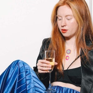 Puede incluir: Una mujer con un blazer negro y una falda plisada azul sostiene una copa de champán. Tiene el pelo rojo y lleva un sujetador negro con pegatinas geométricas de colores en el pecho.