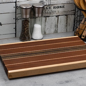 Könnte beinhalten: Rechteckiges Holzschneidebrett mit abwechselnd dunklen und hellbraunen Streifen. Das Brett hat einen hellfarbigen Rand. Salz- und Pfefferstreuer, Holzutensilien und eine kleine Topfpflanze sind im Hintergrund zu sehen.