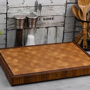 Könnte beinhalten: Ein rechteckiges Holzschneidebrett mit einer hellbraunen Stirnholzoberfläche und einem dunkleren braunen Rand. Das Brett ist etwa 40 cm lang und 30 cm breit. Links steht eine kleine grüne Pflanze in einem weißen Topf.