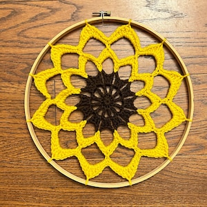 Può includere: Un arazzo da parete a forma di girasole lavorato a uncinetto con un centro marrone e petali gialli. Il girasole è montato su un cerchio di legno.