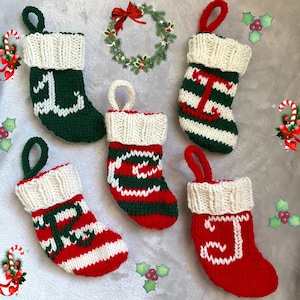 May include: Five knitted Christmas stockings with white cuffs and red and green stripes. Each stocking has a different letter embroidered on the front: J, T, E, C, and I.