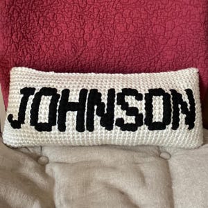 May include: A rectangular, white crocheted pillow with the name "JOHNSON" in bold, black letters. The pillow is resting on a beige surface, with a red quilted background.
