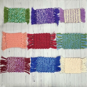 May include: Nine handmade crochet coasters in various colors, including green, purple, peach, red, blue, and cream. Each coaster features a rectangular shape with fringed edges, showcasing a textured stitch pattern. The coasters are arranged on a light wooden surface.