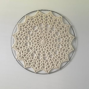 May include: A white crocheted mandala hanging on a white wall. The mandala is circular and has a delicate, intricate design. It is attached to a metal hoop.