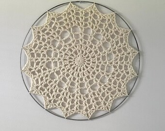 May include: A white crocheted mandala hanging on a white wall. The mandala is circular and has a delicate, intricate design. It is attached to a metal hoop.