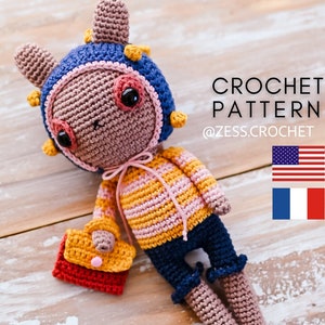May include: Crocheted amigurumi toy bunny wearing a blue and yellow striped hat, a pink and yellow striped sweater, blue jeans, and holding a red and yellow purse. The toy is laying on a wooden surface. The text "CROCHET PATTERN @ZESS.CROCHET" is visible in the image. The American and French flags are also visible in the image.