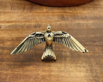 May include: A detailed brass bird figurine with outstretched wings, showcasing intricate feather detailing. The bird is positioned on a wooden surface, with a circular wooden element in the background. The figurine's colour is a warm gold.