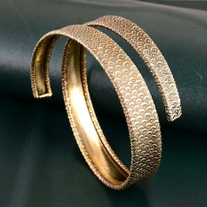 May include: A pair of gold-tone metal bangles with a textured, scaly design. The bangles are open and can be adjusted to fit different wrist sizes.