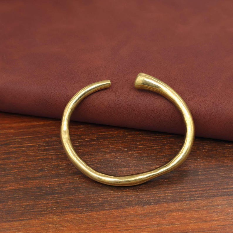 May include: A gold-colored, open-ended cuff bracelet. The bracelet has a smooth, rounded design with a slight hammered texture. The open ends allow for easy wear. The bracelet is displayed on a wooden surface.