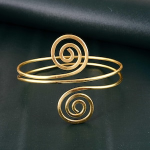 Puede incluir: Un brazalete de metal dorado con un diseño de doble espiral. El brazalete está hecho de dos bandas delgadas de metal que se retuercen juntas para crear una forma de espiral. El diseño de espiral se repite en ambos extremos del brazalete.