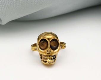 May include: A gold-coloured skull ring. The ring features a detailed skull design with large eye sockets, a prominent nose, and a detailed jawline. The ring band has a textured design. The ring is set against a white background.