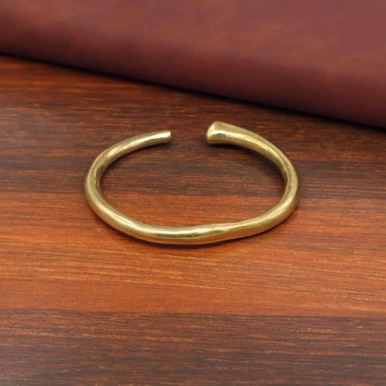 May include: A gold-colored, open-ended cuff bracelet. The bracelet has a smooth, slightly irregular surface and a rounded shape. The ends are slightly thicker and rounded. The bracelet is displayed on a dark wood surface.