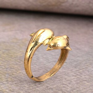 Op de afbeelding: Een ring van goudkleurige metaal met twee in elkaar verweven dolfijnen, een naar voren en de andere naar achteren gericht. De dolfijnen zijn gedetailleerd en hebben een gladde, gepolijste afwerking.
