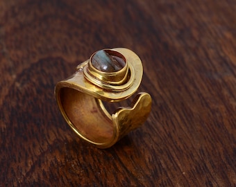 May include: A gold-colored ring with a large, oval-shaped stone. The ring has a unique, asymmetrical design with a hammered texture and a swirling accent. The stone is a mix of brown, gray, and blue hues, set in a raised bezel.