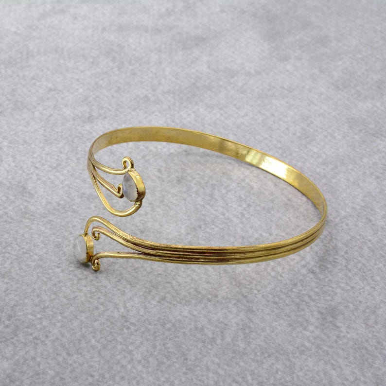 May include: A gold-toned bangle bracelet with two white oval-shaped stones. The bracelet is open and has a delicate, intricate design.