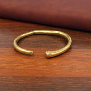 May include: A gold-colored, open-ended cuff bracelet. The bracelet has a slightly irregular, organic shape. The metal has a warm, polished finish. The bracelet is displayed on a wooden surface.
