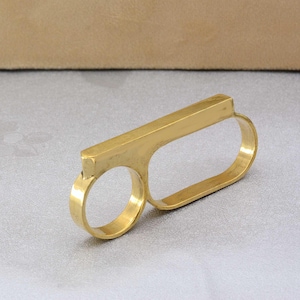 May include: A gold-toned double finger ring with a rectangular bar design. The ring is made of metal and has a smooth finish.