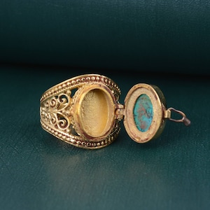 May include: A gold-colored ring with an open locket. The ring has an ornate filigree design and a turquoise stone inside the locket. The ring is on a green surface.