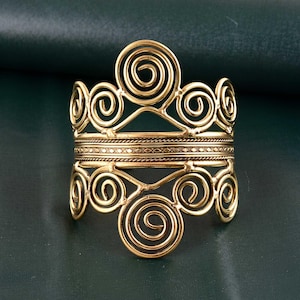 May include: A gold-toned metal bracelet with a decorative design of interlocking spirals. The bracelet has a wide band and is made of multiple layers of metal.