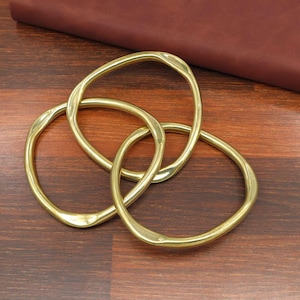 May include: Three intertwined, oval-shaped brass rings. The rings have a smooth, polished surface and a warm, golden hue. They are arranged on a dark wooden surface, creating a visually appealing geometric pattern. The rings are a decorative element.