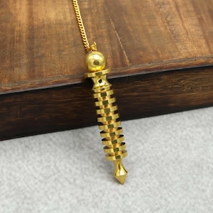 Puede incluir: Un péndulo dorado con una bola en la parte superior y una serie de discos circulares apilados que forman el cuerpo. El péndulo cuelga de una delicada cadena dorada. El fondo es una superficie de madera.