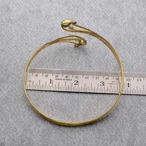 May include: A gold-toned metal bangle bracelet with a decorative design featuring two swirls and a small round accent. The bracelet is measured against a ruler.