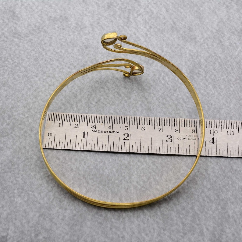 May include: A gold-toned metal bangle bracelet with a decorative design featuring two swirls and a small round accent. The bracelet is measured against a ruler.