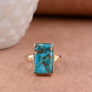 May include: A gold-colored ring featuring a rectangular turquoise stone with brown matrix. The stone is set in a gold bezel and has a polished finish. The ring's band is also gold-colored.