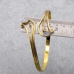 May include: A gold-toned bangle bracelet with two white moonstone accents. The bracelet is measured against a ruler, showing it is approximately 5 inches in diameter.