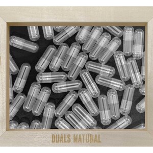 May include: A light-colored wooden frame containing a collection of empty, clear, two-piece gelatin capsules scattered over a black background. The frame features the text NYC on the left, 1989 on the right, and DUALS NATURAL on the bottom.
