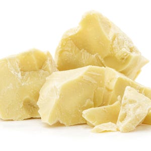 May include: Several irregular, solid chunks of pale yellow cocoa butter. The pieces have a waxy, matte texture with sharp, jagged edges and varying sizes, stacked together to form a cluster of raw, unrefined vegetable fat.