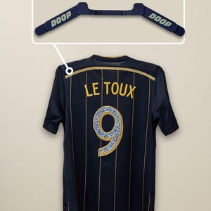 May include: Navy blue soccer jersey with gold vertical stripes and the name "LE TOUX" and the number "9" on the back. A blue hanger with the word "DOOP" is above the jersey.