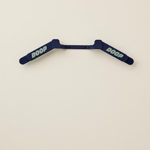 May include: A navy blue plastic hanger with the word "DOOP" printed in light blue on each end. The hanger is mounted on a white wall.