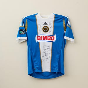 May include: Blue and white soccer jersey with gold accents, featuring the Adidas logo and the word "BIMBO" in red. The jersey has the MLS logo on the sleeve and is covered in signatures.