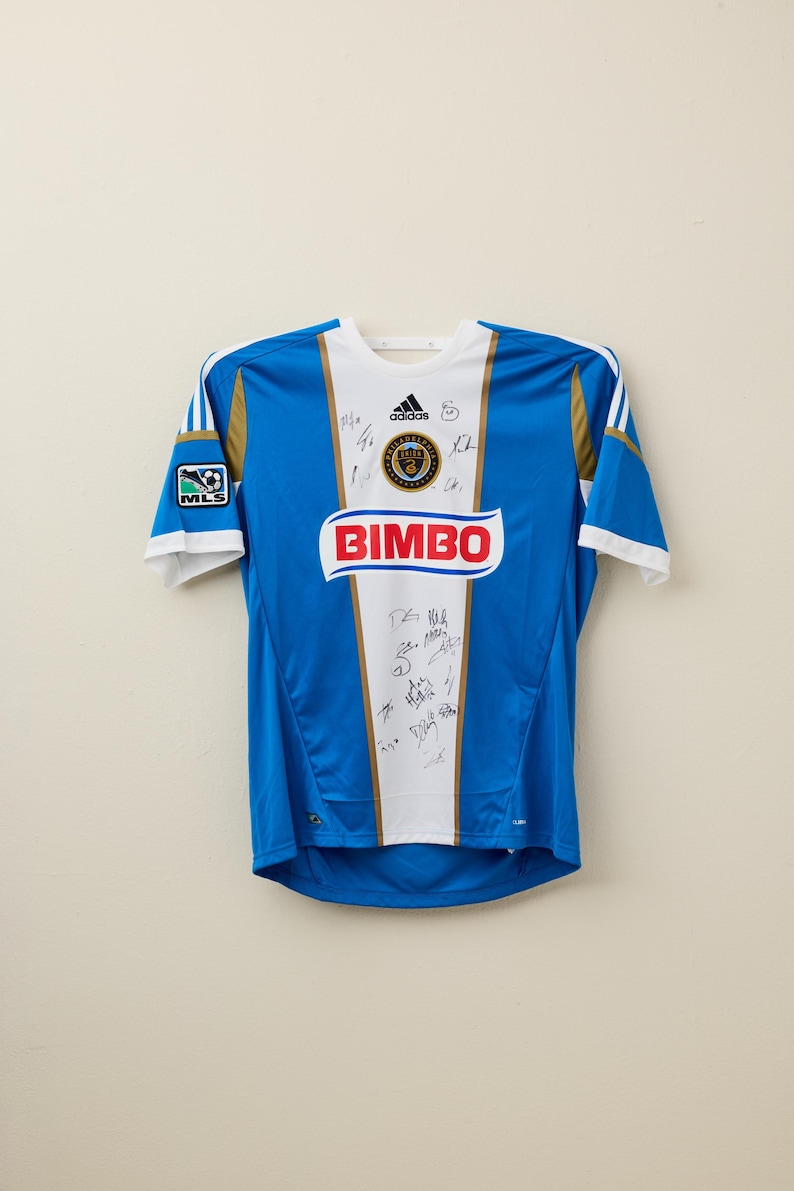 May include: Blue and white soccer jersey with gold accents, featuring the Adidas logo and the word "BIMBO" in red. The jersey has the MLS logo on the sleeve and is covered in signatures.