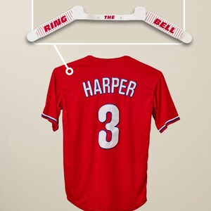 May include: A red baseball jersey with the name "HARPER" and the number "3" in white. Above the jersey is a white hanger with the words "RING THE BELL" in red.